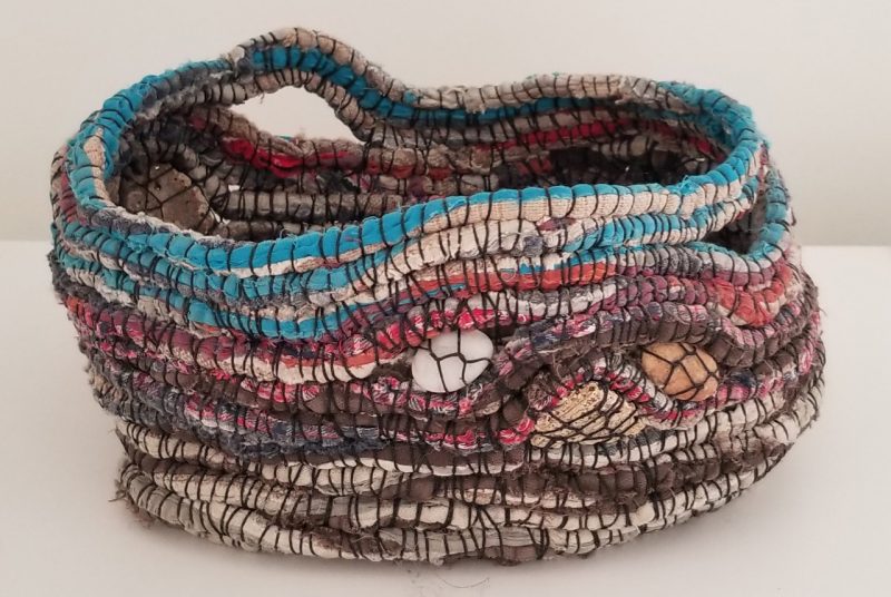 This relaxed-looking, unstructured basket by artists Martha Olson is brown and beige-colored with accents of red and turquoise. It was made with rag rug and found stones. 
