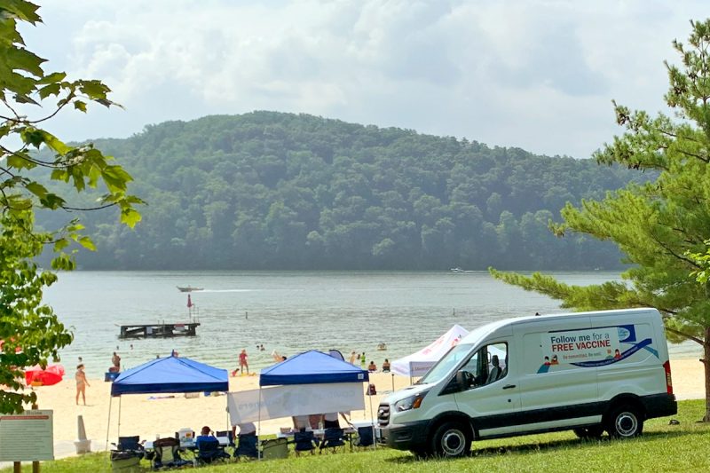 A mobile vaccine clinic , complete with a van, chairs, tents, and tables, is set up at Claytor Lake State Park . In the distance, there is a man-made beach and water. 