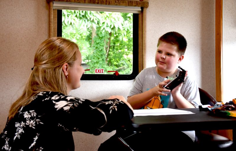 Graduate student clinician Jennifer Bertollo works with Cory, a young client, inside the Mobile Autism Clinic RV. Cory sits at a table with a puzzle toy in hand. Jennifer is kneeling to be at eye level with Cory.