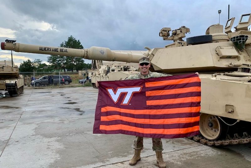 Hokie Heroes 2021 U.S. Army Capt. Daniel Gaines holds a Virginia Tech flag in front of a tank.