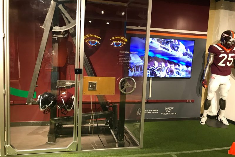 An exhibit at the Science Museum of Western Virginia includes a mannequin dressed in a Virginia Tech football uniform, a screen showing an image of many helmets, and a glass room where two helmets are pushed together using metal arms.