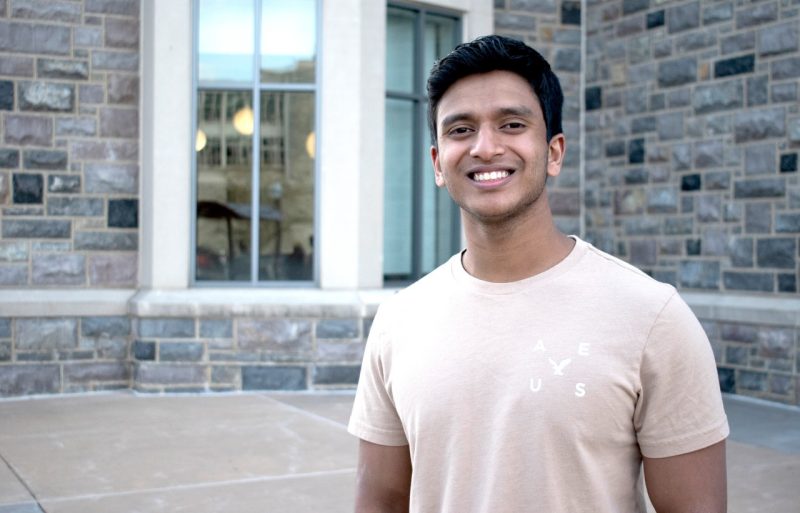 Saket Bikmal, wearing a beige t-shirt, smiles as he poses by the New Classroom Building. Photo for Virginia Tech by Steven Mackay.