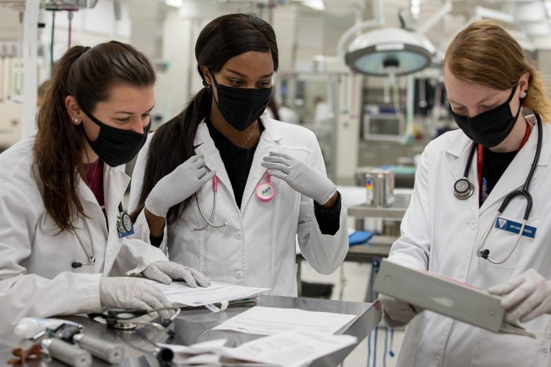 Three veterinary students in white coats and gloves stand around a table.