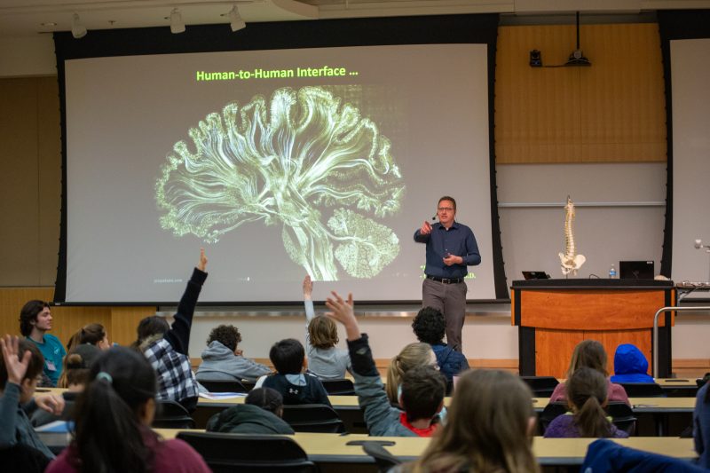 A man stands in front of an audience of children. On the projector screen behind him, there is a giant brain.