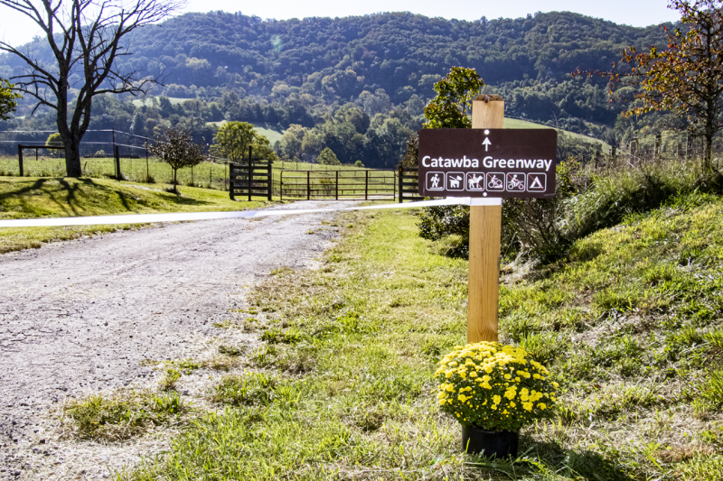 A brown wooden Catawba Greenway sign sits next to a trail that leads off to the mountains beyond.