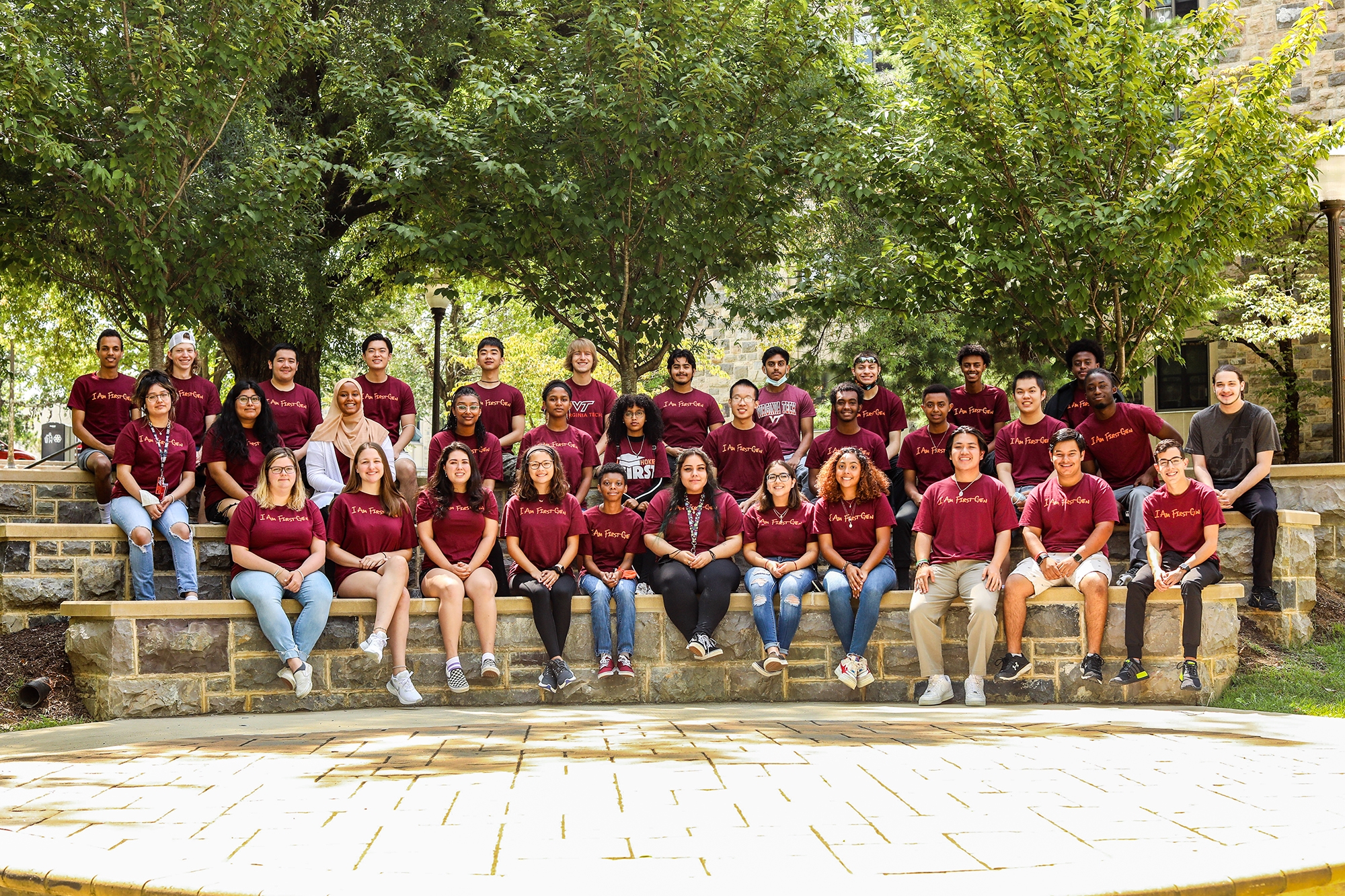 A group of first-generation students sit together on a rock wall outside on campus, posing for a group photo. They are wearing maroon shirts with orange lettering that says, "I am First Gen."