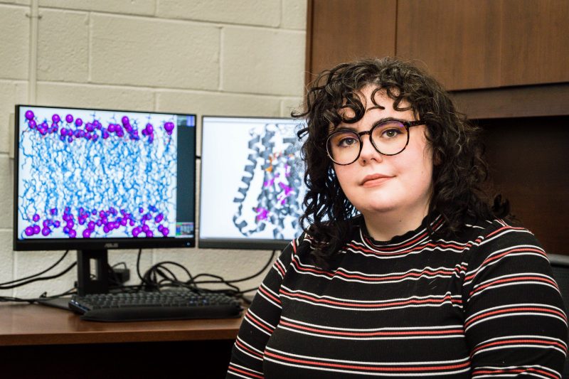 Julia Montgomery, Virginia Tech Biochemistry Graduate Student sits in front of computers with two protein structure computations