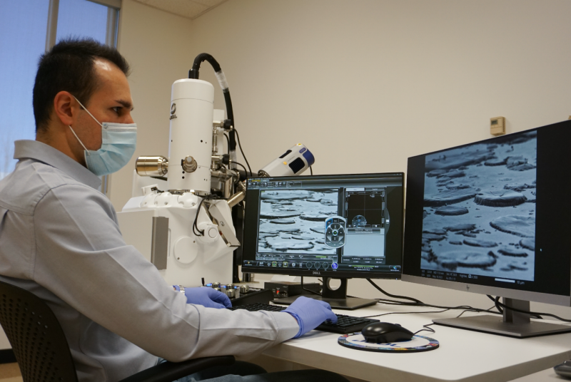 A person in a laboratory looks at images displayed on a computer screen beside a piece of equipment.