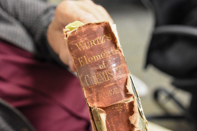 Jay Stipes holds a well-worn, historic copy of Elements of Chemistry by Charles Adolphe Wurtz. The book belonged to John McLaren McBryde. The title is printed in gold font against a brown, now fading cover.