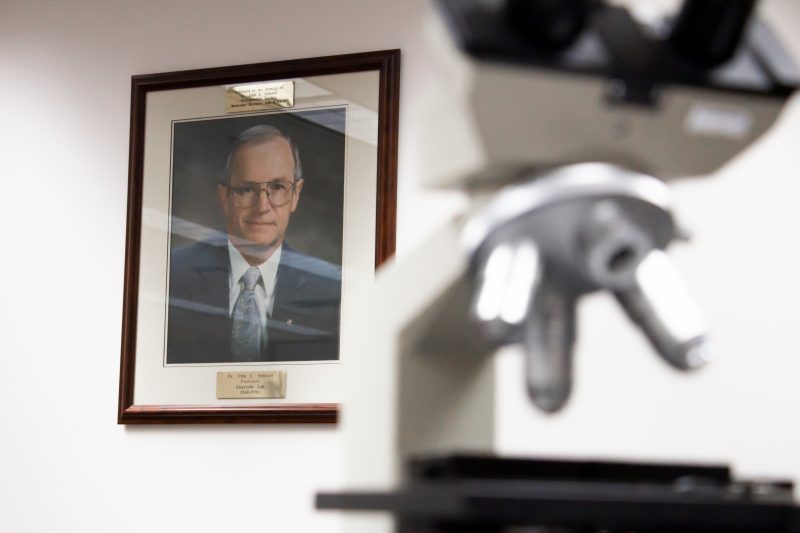 A photograph of John L. Johnson hangs in Teaching Lab 2 in Fralin Hall. Under the photo of the man, there is a caption that reads: "Dr. John L. Johnson. Professor. Anaerobe Lab. 1968-1996." A microscope is blurred out in the foreground. Photo courtesy of Tyler Harris for Virginia Tech.