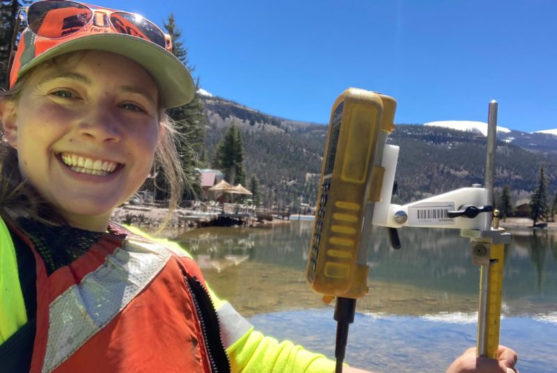 Woman posing for a selfie while standing on the shore of a lake with pine-covered mountains in the background and holding a rod with graduated markings with some type of monitor attached