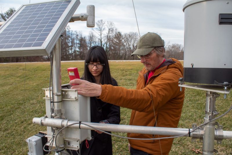 Student and professor examine a weather station composed of a solar panel and various metal boxes, tubes, and a cylinder
