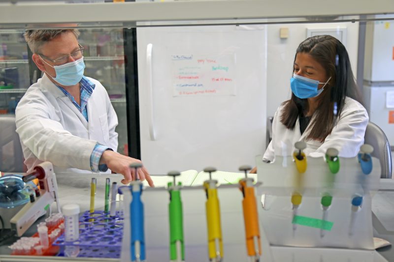 A man and a woman are pointing at a laboratory notebook. Four brightly colored pipettes are in the foreground.