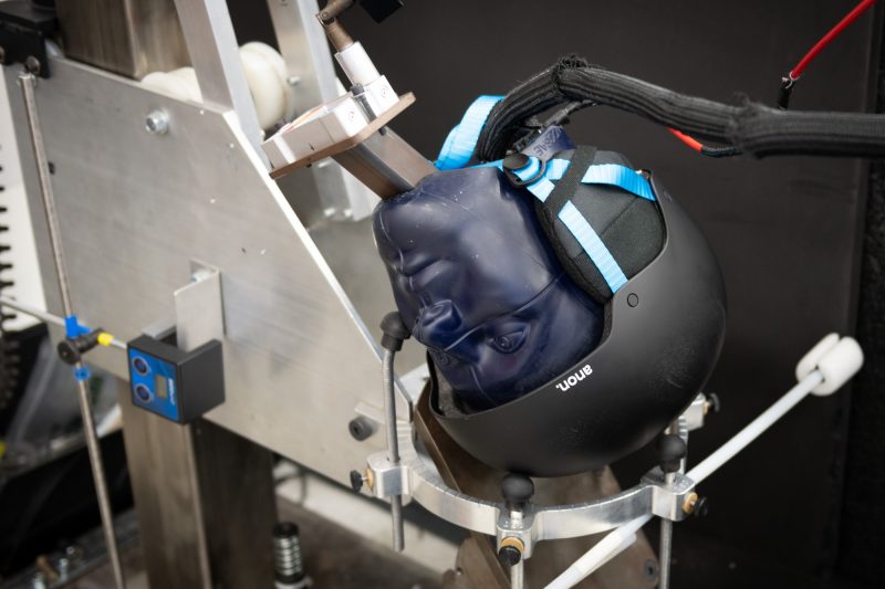 A blue dummy headform wearing a black ski helmet rests on an angled metal anvil 