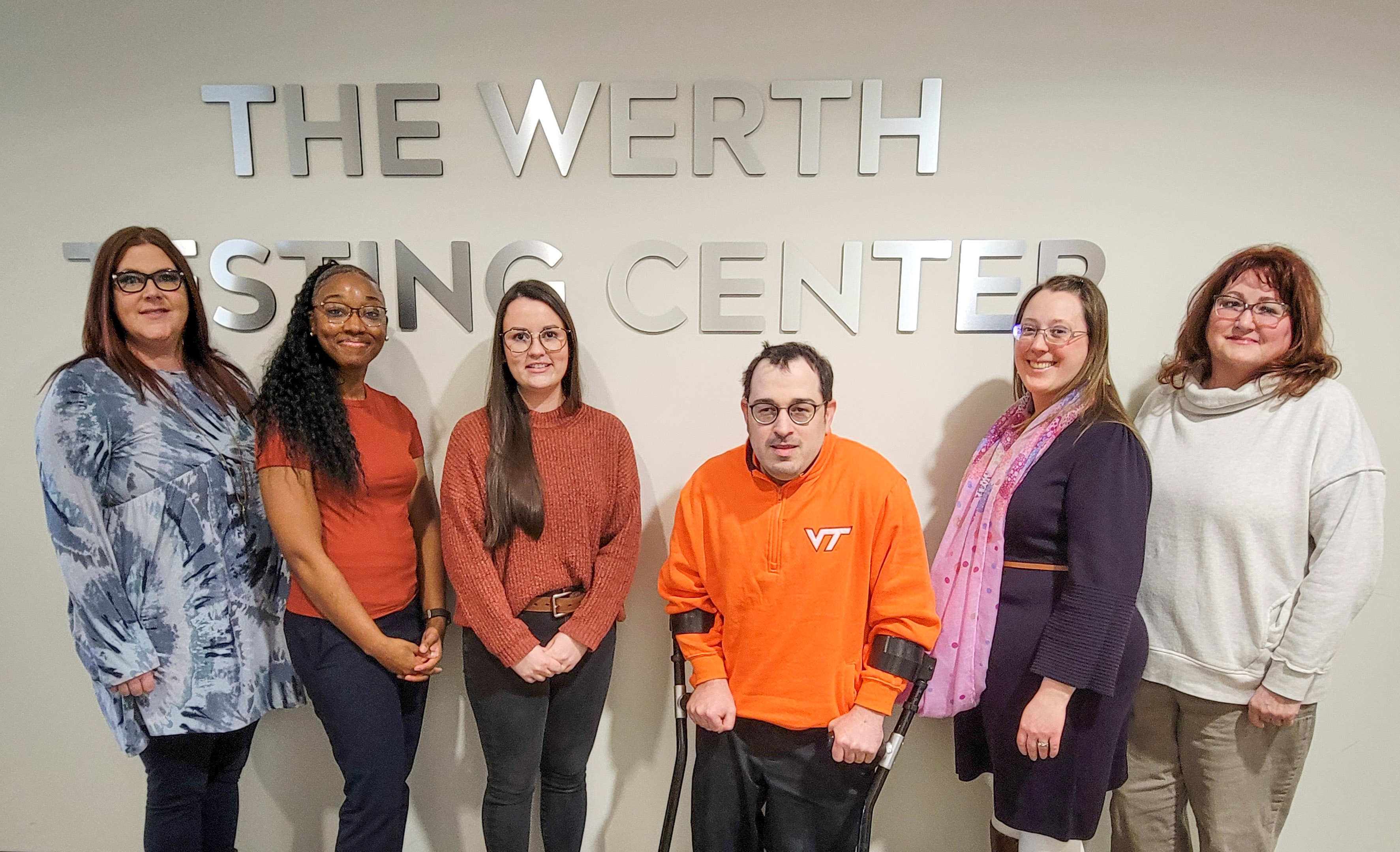 6 individuals stand in front of a silver sign that reads, "The Werth Testing Center." From left to right, there are: A tall, middle-aged white female wearing a blue blouse and shoulder-length auburn hair; a young, black female with dark curly hair, wearing a bright orange top; a young white female with longer brown hair wearing an orange sweater; a short middle-aged white male wearing a Virginia Tech sweatshirt and standing on forearm crutches; a middle-aged white female with shoulder length blonde hair and a purple dress; a middle-aged white female with short red hair and a white blouse. They are all wearing glasses, and smiling for a photo while standing beside one another.
