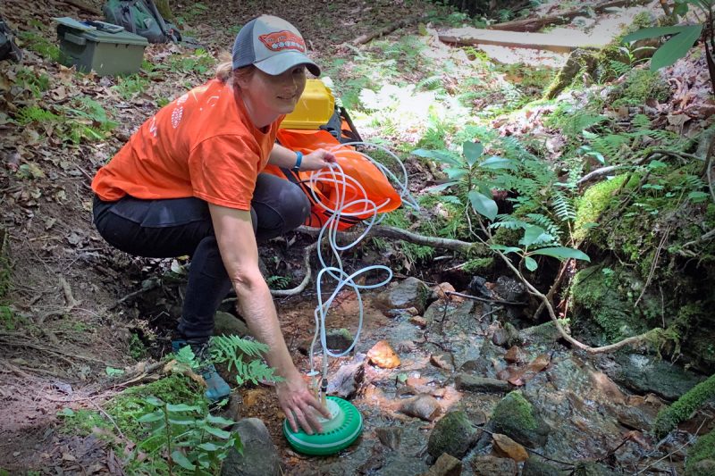 A woman in an orange shirt is measuring greenhouse gases in a stream.
