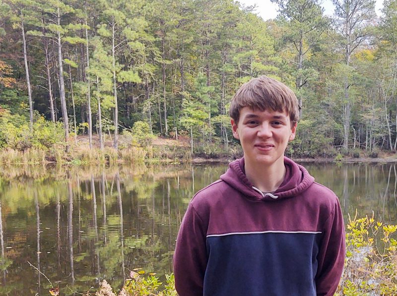 High school junior John Button, in a blue and maroon hoodie, stands in front of a pond surrounded by pines and shrub vegetation on an overcast day.