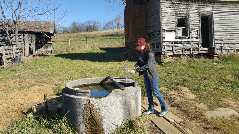 A woman in a maroon Virginia Tech hat leans over a large concrete cylinder that has water inside of it. She is collecting water from a small pipe that is jutting out of it.