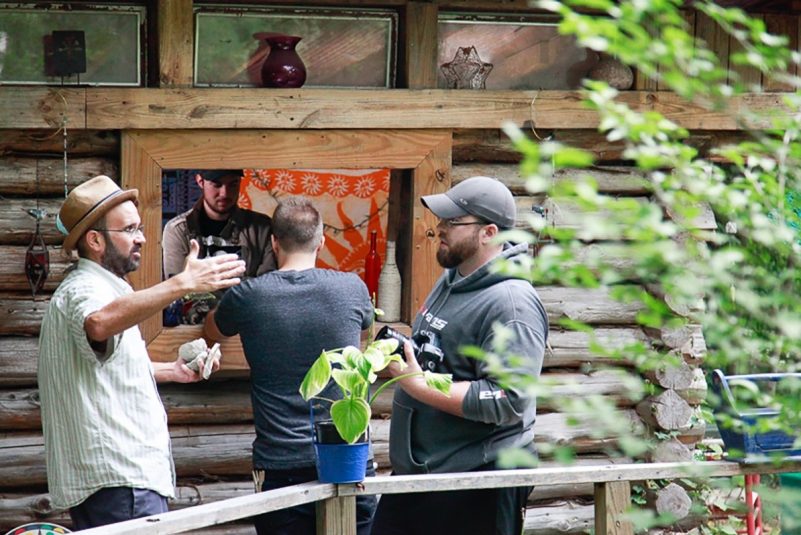 Creator/Executive Producer Chris Valluzzo wears a straw hat and gestures with his hands while he talks with Director of Photography Darren Van Dyke in an outdoor location. A camera operator focuses on equipment behind the two.