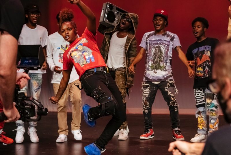 Six Memphis Jookin' dancers gather around one female dancer to encourage her performance. All are dressed in street clothes - t-shirts, ripped jeans, and baseball caps. One member holds a large boombox over his shoulder.