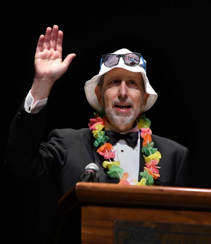 Dean Lee Learman in a tuxedo wearing a lei around his neck, and a bucket hat with sunglasses on top of his head. He had one hand raised in celebration