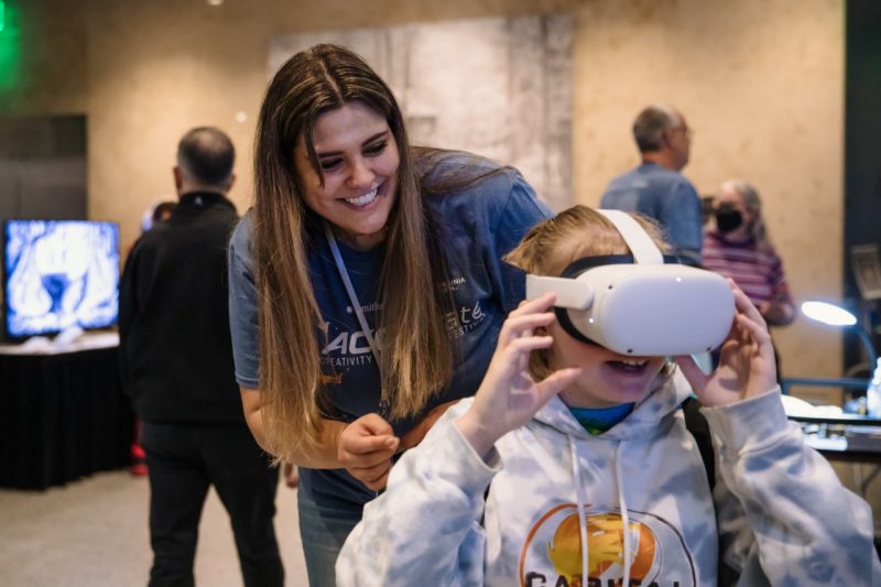 A Virginia Tech student helps a child put on a VR headset at the Benthos 360 exhibit at the Accelerate Festival