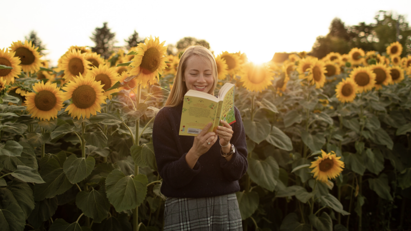 Morgan Long reads a book while standing in a field of sunflowers.