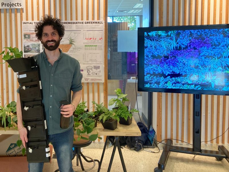 Zachary Gould holding stack of plants with a table of plants behind him and a screen showing an animation of plants + research.