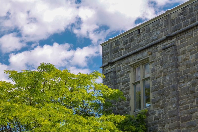 The top of a grey hokie stone building on a summer day surrounded by green trees