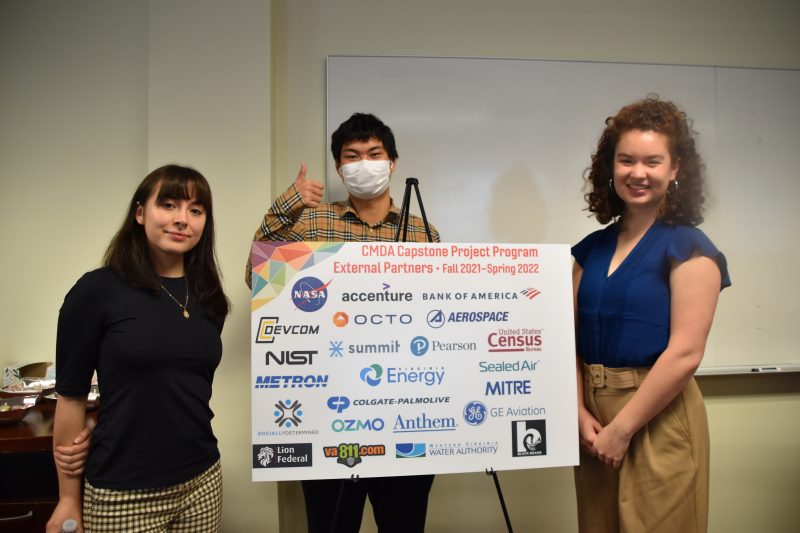 Three Virginia Tech students stand around a sign displaying the logos of the CMDA capstone project program's external partners.
