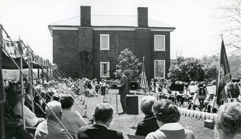 Black and white photo of a crowd sitting outside a brick house.