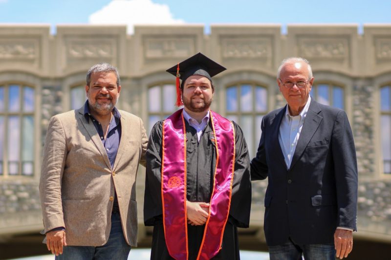 Three people pose for outdoor photo with the center one in a graduation robe