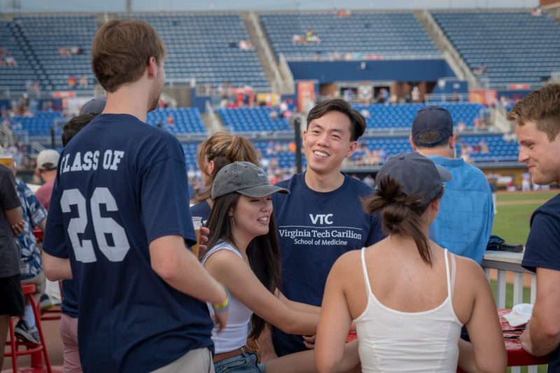 A student wearing a Virginia Tech Carilion School of Medicine t-shirt smiles at another with Class of 26 on the back of his t-shirt at a baseball game. 