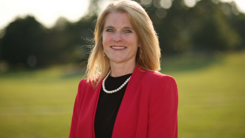 Portrait of Robin Queen with grassy hill and dark green trees in background. She wears a red blazer, black shirt, and pearls.