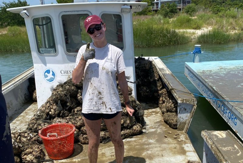 Kylie Harris stands on the deck of a boat with mesh bags of oysters on the desk behind her.