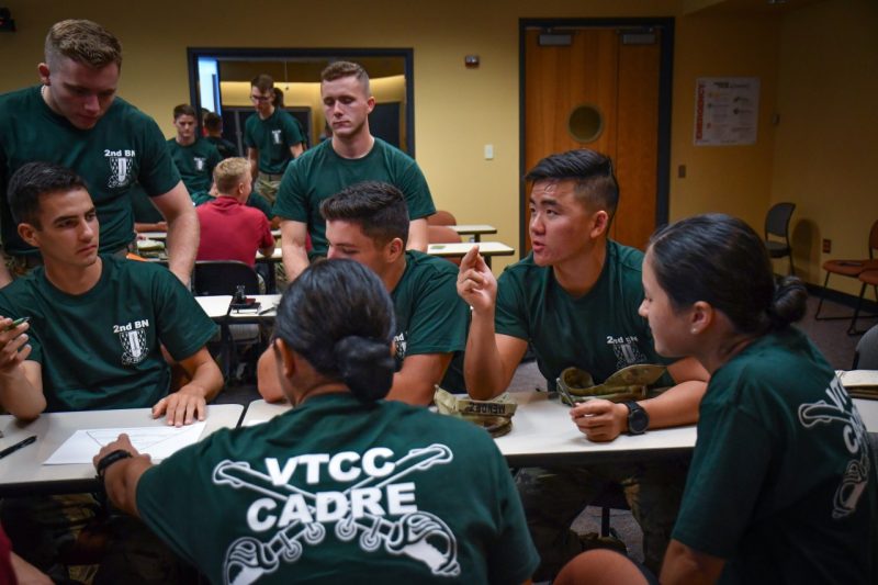 In a classroom setting, a group of seven cadets in green VTCC Cadre shirts face each other discussing sexual assault prevention. Other cadets can been seen in the background also discussing the topic.
