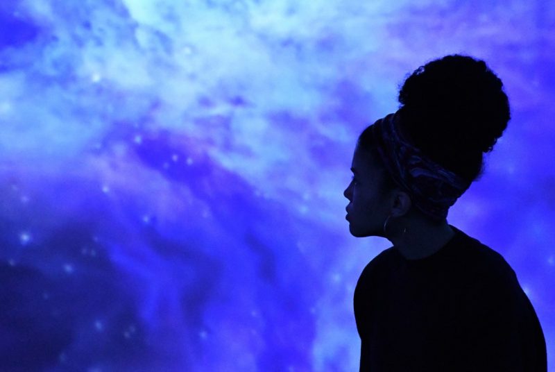 A woman in shadow sits in front of a screen that looks like a scene from space, filled with blue and purple swirls and what looks like stars. 