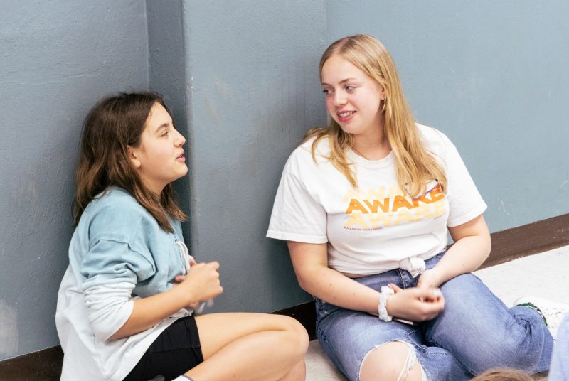 A dark-haired middle school girl has a conversation with a blonde Virginia Tech mentor while sitting on the floor of a room in Squires.