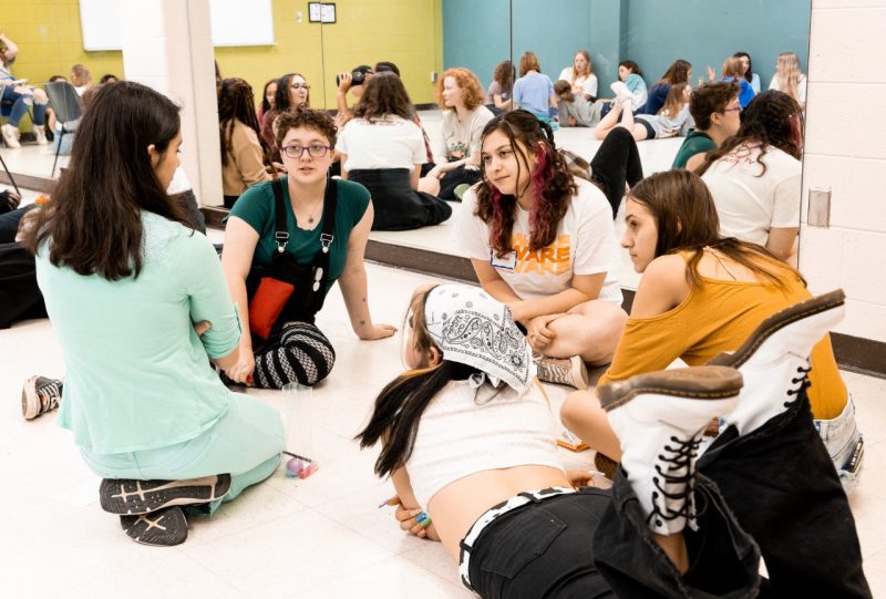 A Virginia Tech student in an AWARE mentor sits on the floor in a group with four middle school girls, listening intently to one with dark hair