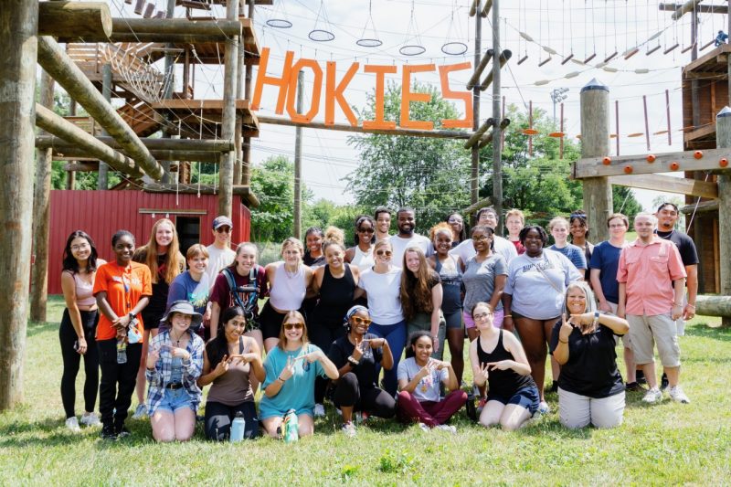 A diverse group of college students pose in front of the ladders and ropes of a high-ropes challenge course at Virginia Tech.