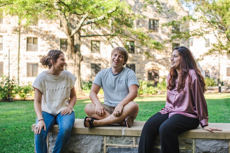 Three Residential Well-being initiative student leaders sit on a Hokie Stone terrace and talk, outside on a summer day. 