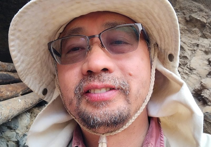 A man with a salt and pepper goatee and large white hat looks into his own cell phone camera. 
