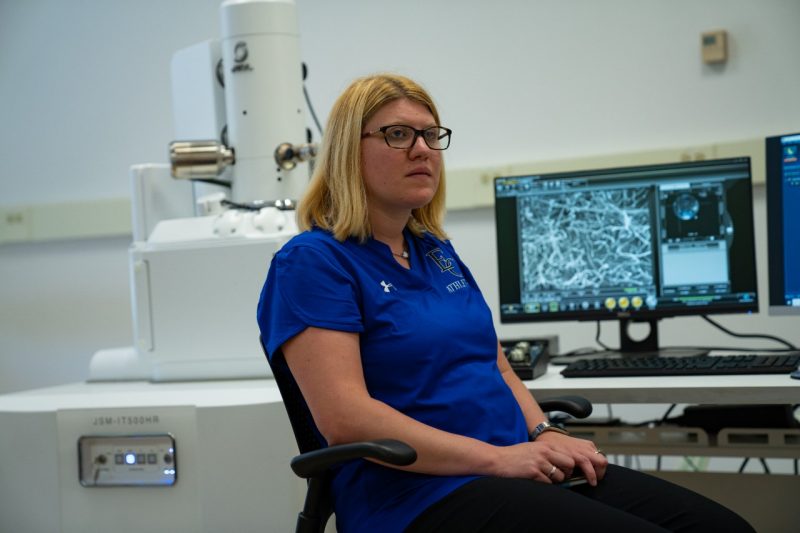 A woman in a blue polo sits in front of a computer screen with an image of a electron microscopic enlargement on it.
