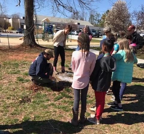 group of people stand around a man crouching down as he holds a small plant