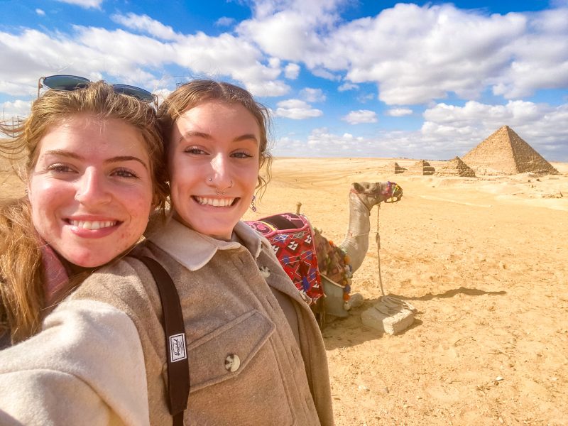Two students taking a selfie in front of a camel and a pyramid
