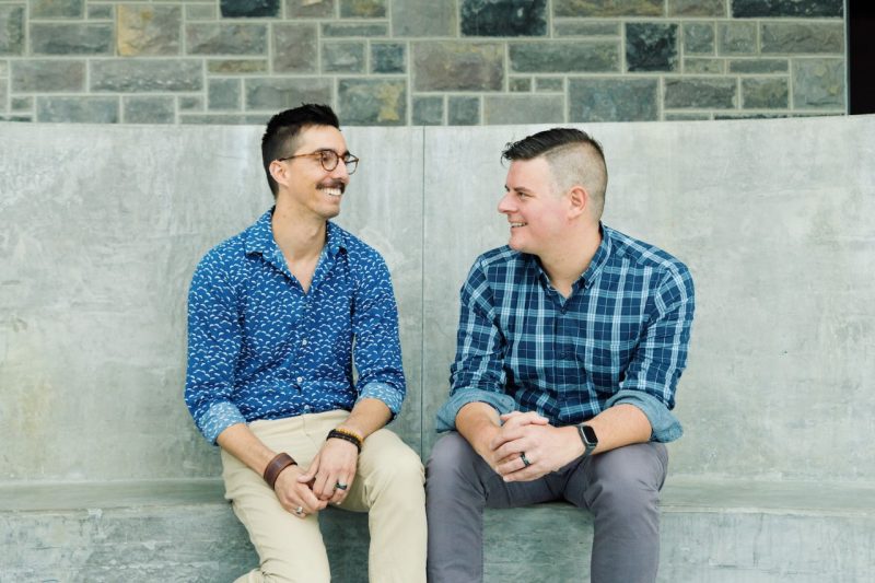 Husbands Chris Campo Bowen, in a blue shirt, and Michael Robert, in a plaid shirt, laugh together on a cement bench.