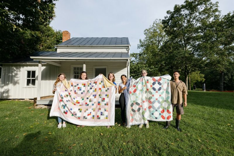 Six students stand in the lush grass outside Fraction House to show off two tied quilts, one in shades of pink and red and yellow, the other circles of red and aqua and green.