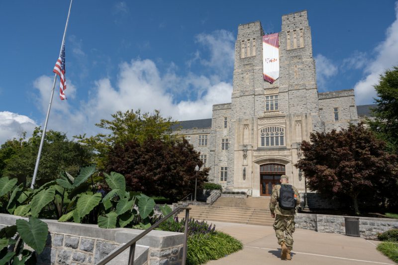 A member of the Virginia Tech Corps of Cadets walks up the grey steps to grey Hokie Stone Burruss Hall on a sunny day