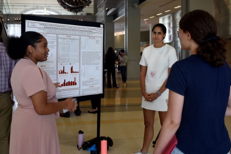 Two students stand in front of a presentation poster, describing their research results to another student.