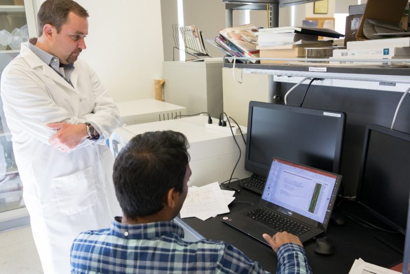 Scott Verbridge, in a white lab coat, stands while Barath -- in a blue plaid shirt -- sits and they analyze research results on a laptop screen.